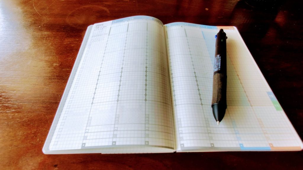 ジブン手帳mini 2026を開いてペンを添えた使用イメージ（木目背景）